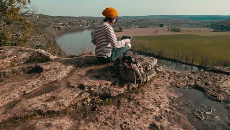 A-woman-opens-the-screen-of-her-laptop-on-the-edge-of-a-cliff,-a-camera-is-placed-on-her-backpack