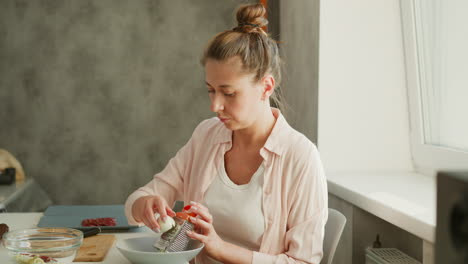 female chef focused on grating cheese into bowl while preparing meal in bright kitchen, seated at white table with cutting board and transparent glass bowl nearby, creating clean, organized workspace