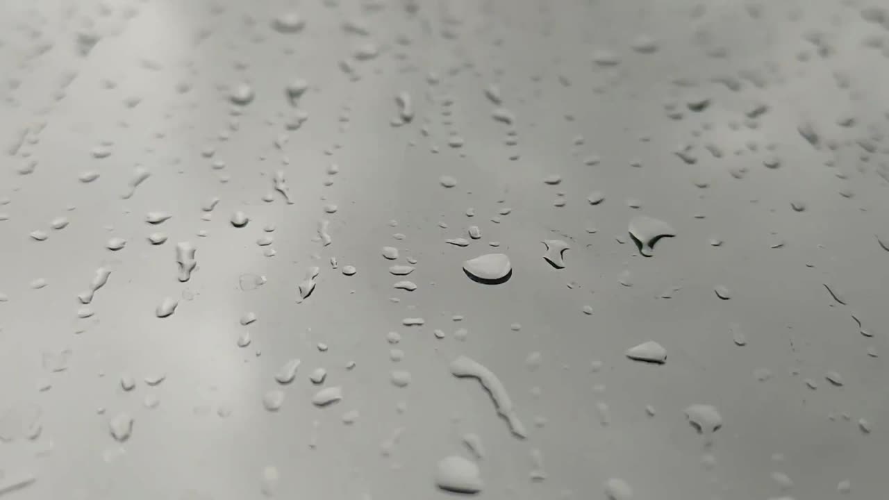 Raindrops fall gradually down a vertical glass surface, guided by gravity. Their slow descent captures the essence of rain's delicate movement.