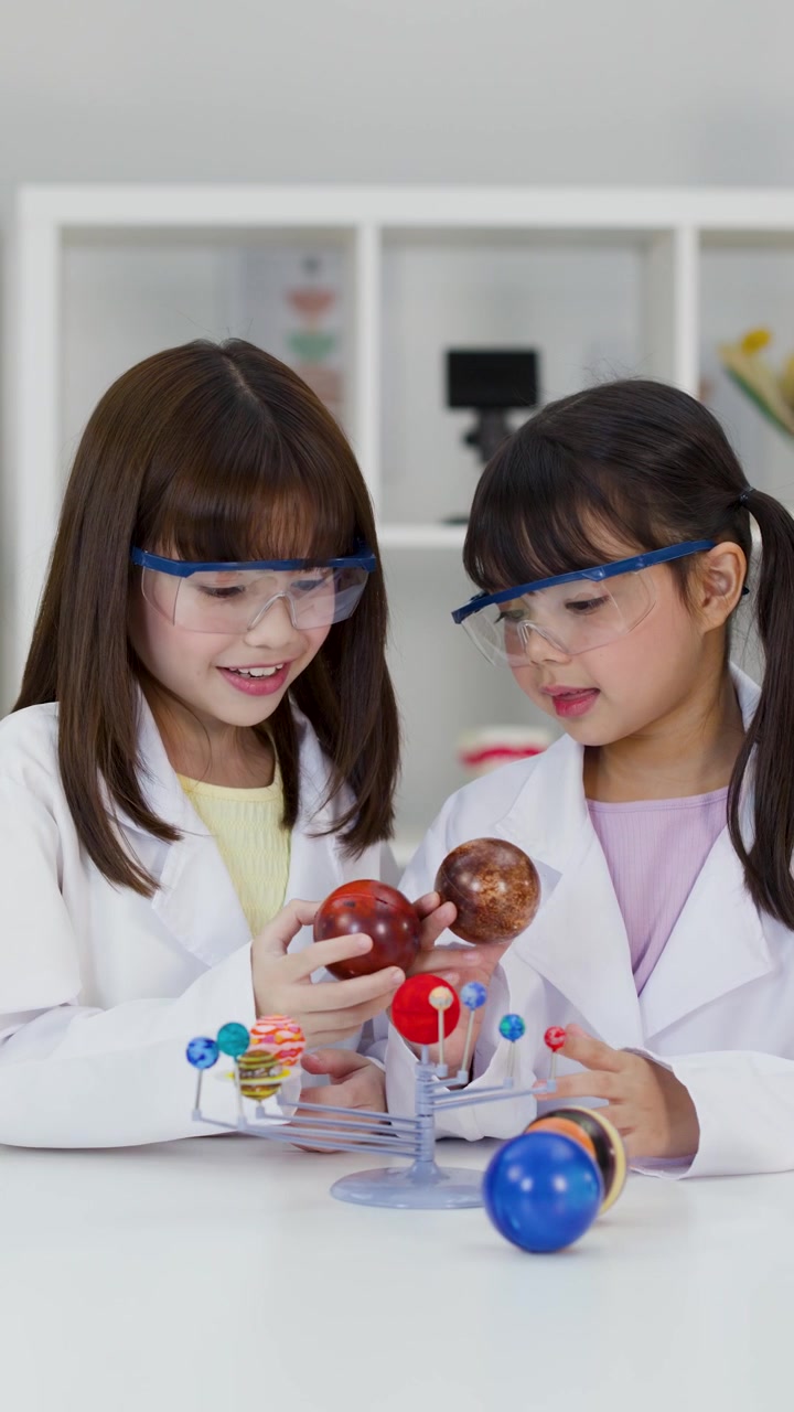 Dua gadis muda mengenakan jas lab dan kacamata pelindung memeriksa model planet di ruang kelas