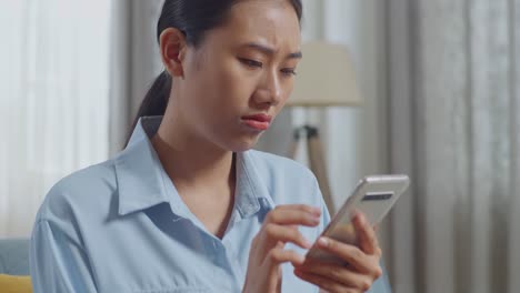 close up of upset asian woman using smartphone while sitting on sofa in the living room