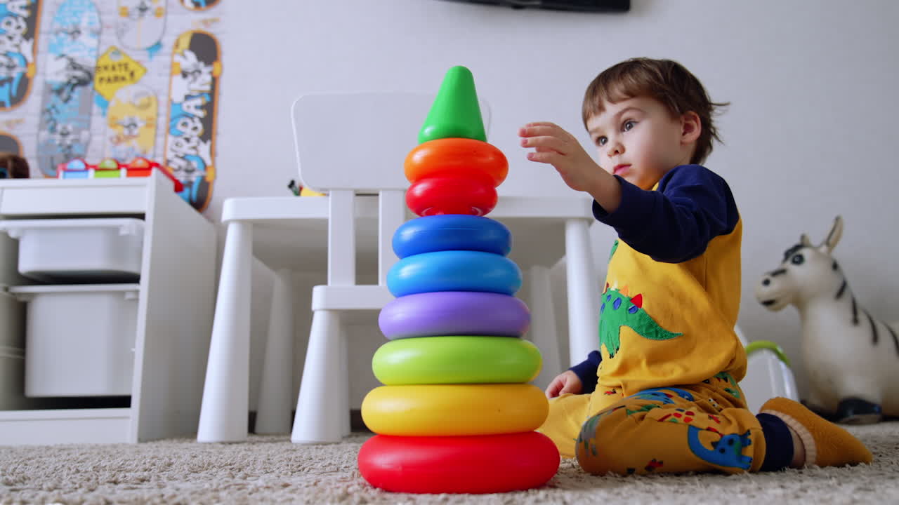 Adorable two-year old toddler building a pyramid at home. Baby boy sits on the floor busy with his game. Low angle view.