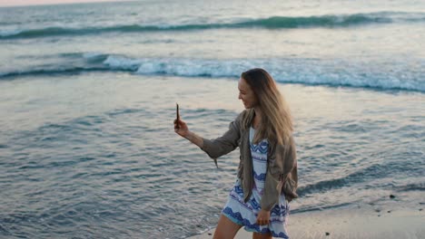 pretty young blonde woman standing on beach during sunset, taking selfie photo and video, spinning dancing and smiling