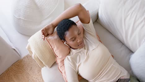 happy african american woman lying on sofa using earphones and listening to music, slow motion