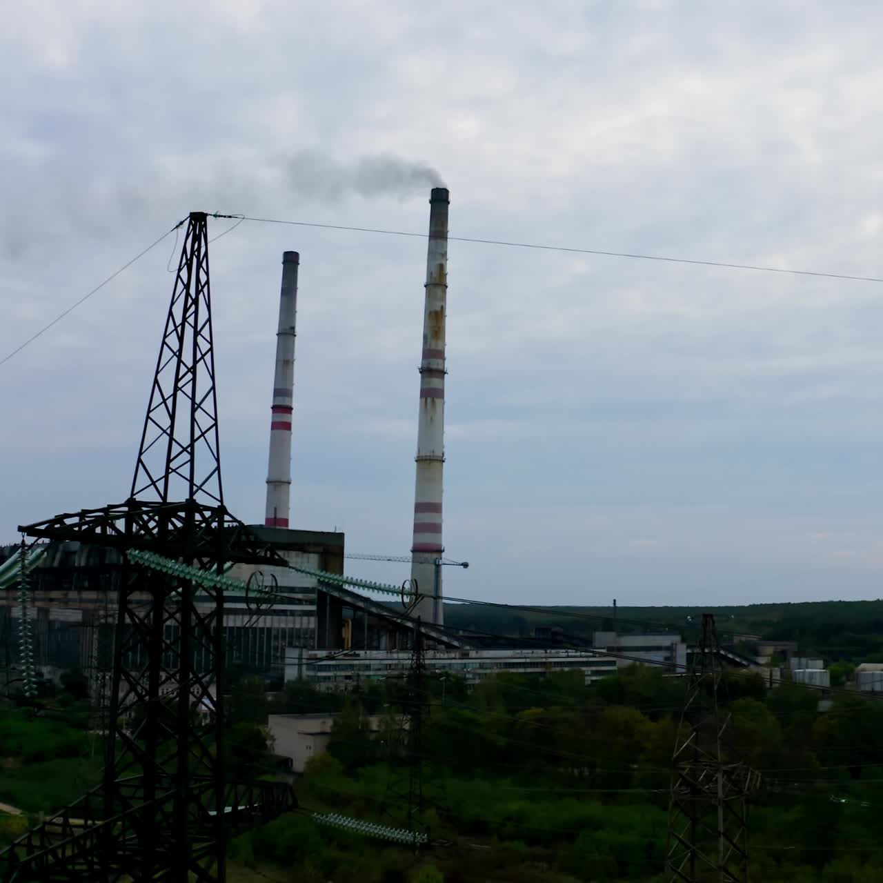 Electricity towers on factory background. High-voltage electric pylons near the harmful enterprise. The concept of nature and technology. Camera rising up.