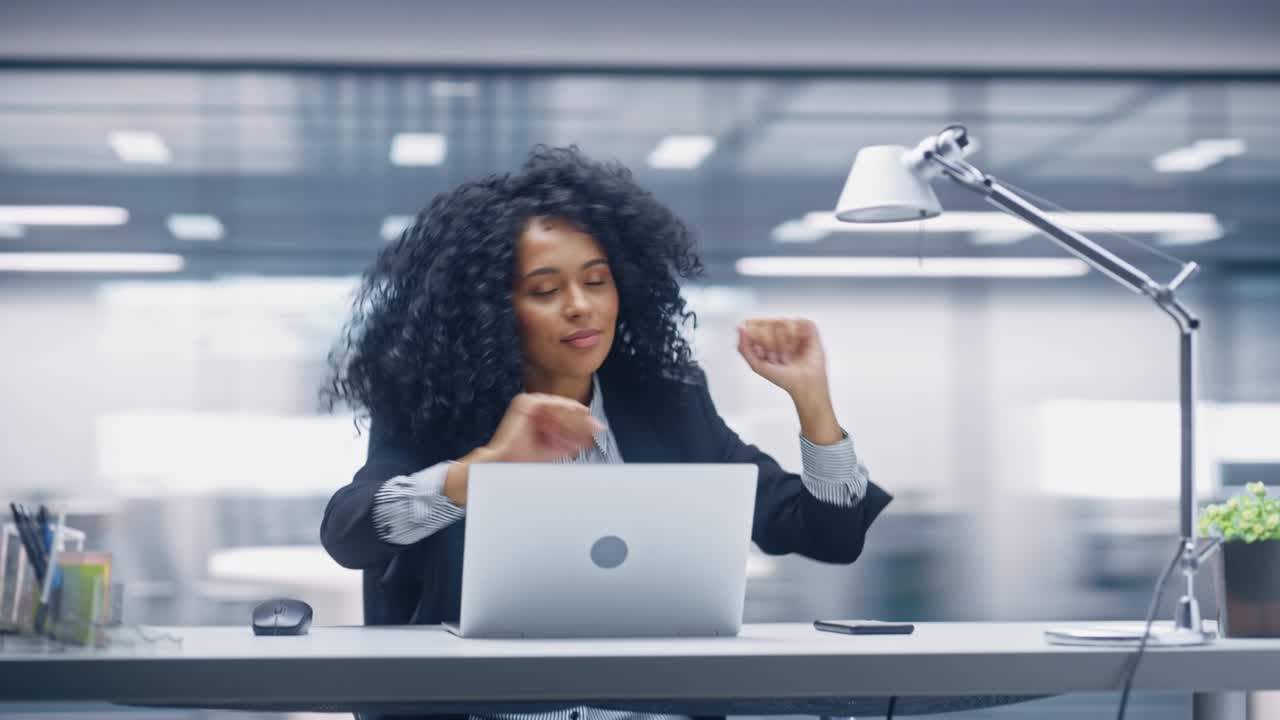 Escritório 360 Graus: Empresária negra dança e sorri enquanto sentada em sua mesa. Bela especialista trabalha no laptop e celebra projeto de e-commerce bem-sucedido. Tomada com câmera em movimento.