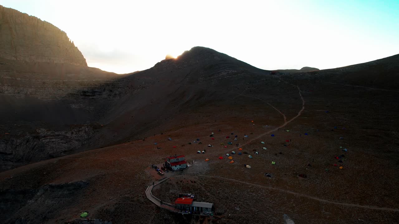 wide aerial shot of a valley under the peak of mountain Olympus while the sun sets and creates lens flares, the shot continues and lowers altitude revealing a camper's hut and lots of colourful tens