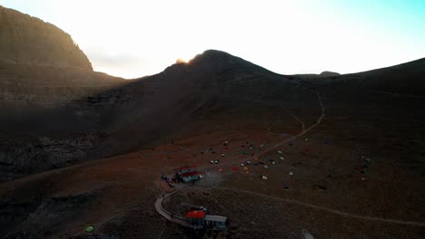 wide aerial shot of a valley under the peak of mountain olympus while the sun sets and creates lens flares, the shot continues and lowers altitude revealing a camper's hut and lots of colourful tens