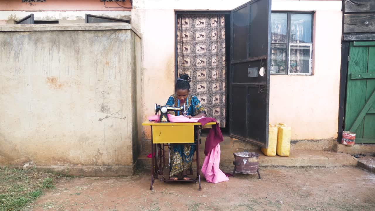 A wide shot of an African woman outside her small house sewing clothing on a manual tailoring machine