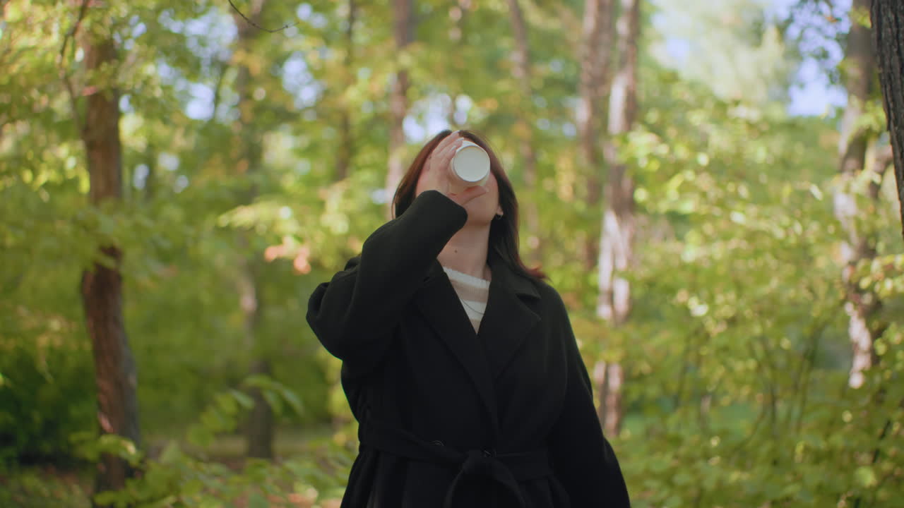 Carefree woman strolls through arboretum holding mug, sipping warm drink, lifting leaf to nose, enjoying forest scent under soft sunlight, calm pause during quiet autumn walk