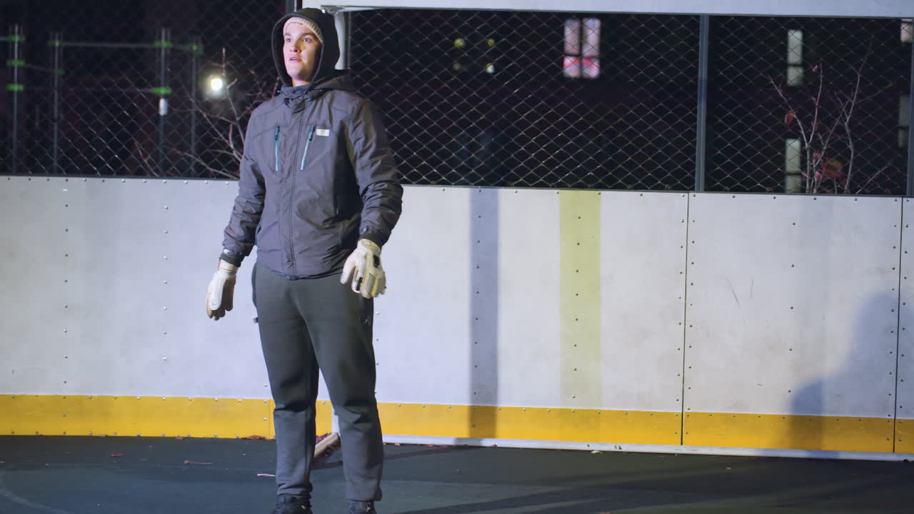 골키퍼는 도시 야외 스포츠 코트에서 야간 훈련 중에 정확하고 집중적으로 축구 공을 필드에 니다. 배경에는 금속 울타리와 조명 된 주거 건물이 포함됩니다.