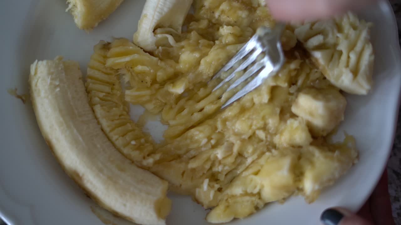 primer plano de arriba de la mano mushing banana con un tenedor en un tazón blanco