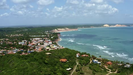 left trucking extreme wide aerial drone landscape shot of the famous tropical tourist beach town of pipa, brazil in rio grande do norte with small waves, cliffs, golden sand, and green foliage