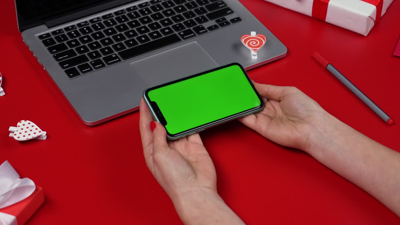 여성의 손은 초록색 화면 크로마 키를 가진 스마트폰을 수평 위치로 들고 있습니다. 노트북, 선물, 심장 및 발렌타인과 함께 빨간 테이블의 상단 뷰. 발렌타인의 날 개념. 근접. 느린 모션.