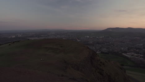 Panning-shot,-cityscape-overlooking-the-whole-city-of-Edinburgh-moments-after-sunset-from-Arthurs-Seat-with-wonderful-blue-hour-light