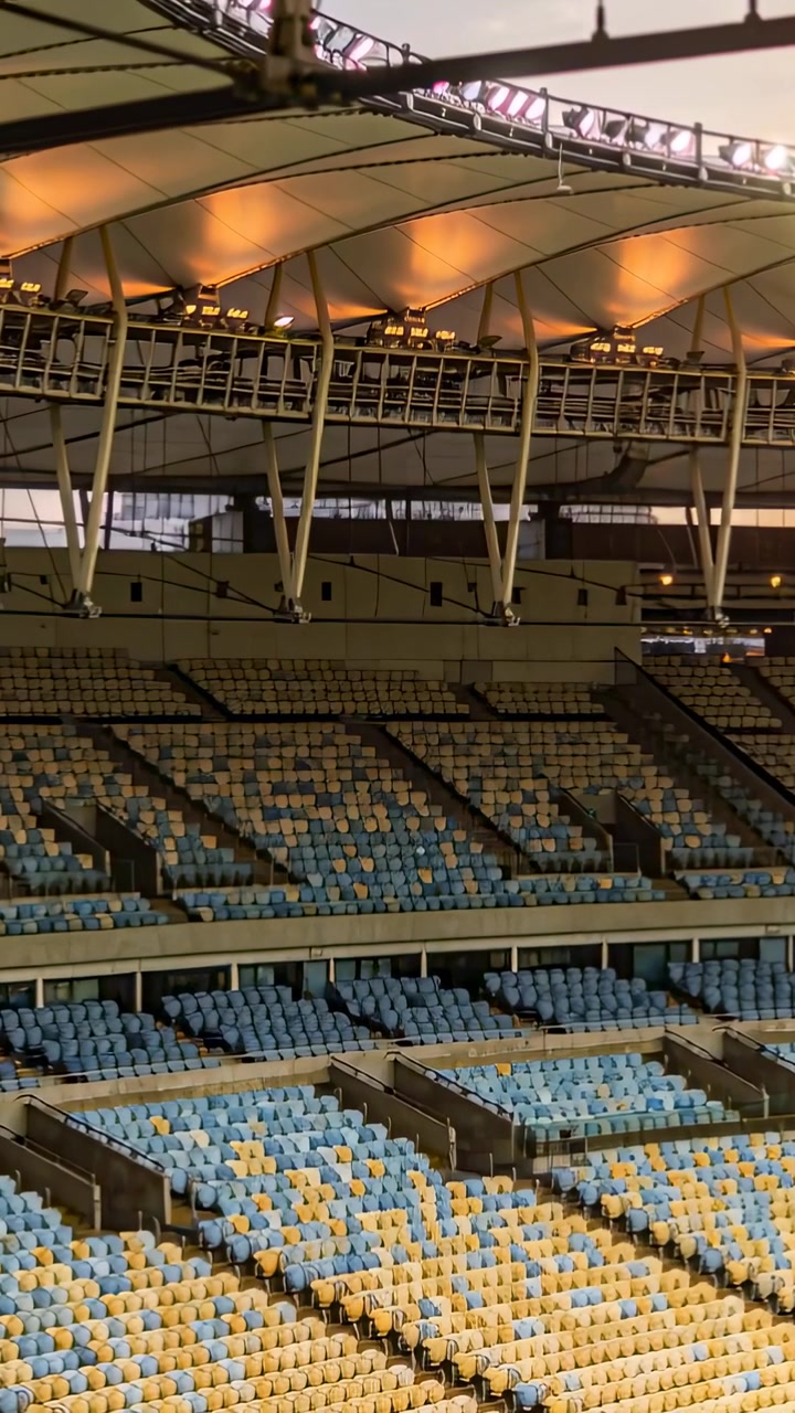 Vertical video: Panning shot revealing empty blue-yellow seats under canopy at stadium, lit soffits