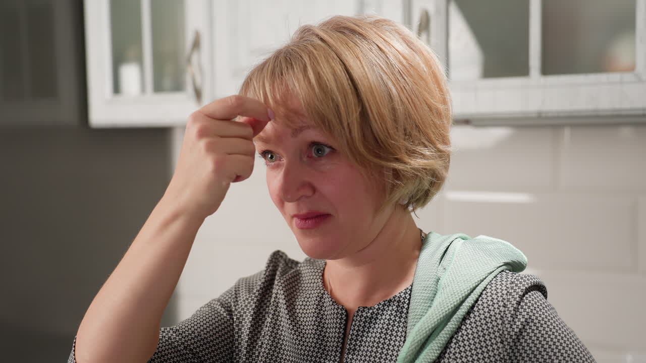 Housewife with towel on shoulder gently touches fringe hairstyle while adjusting hair in invisible mirror, expressing subtle confidence and care inside bright kitchen space with white tiled cabinets