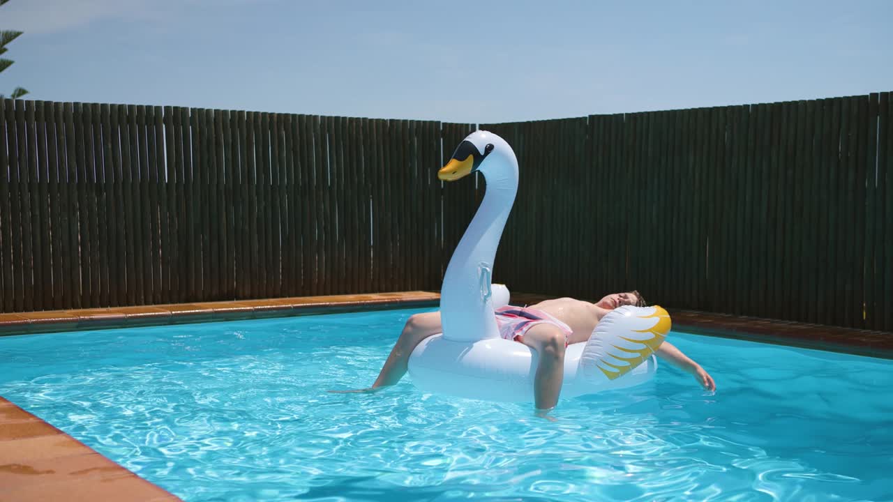 Young happy boy relaxes and sunbathe on inflatable swan toy in blue pool.