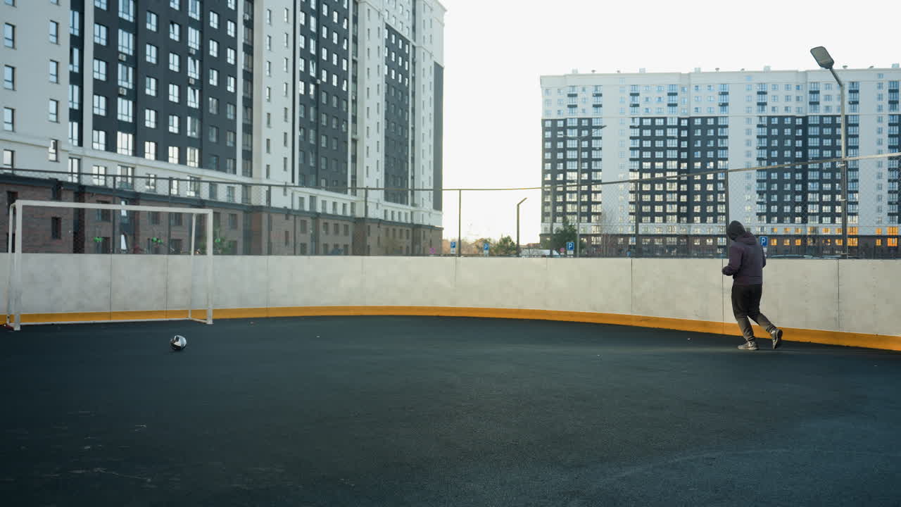 amplia vista de un deportista corriendo en una arena al aire libre con una pelota de fútbol y un poste de meta con edificios urbanos de gran altura en el fondo