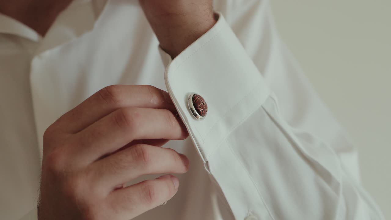 Close up of groom fastening cufflink on white shirt sleeve during wedding preparation
