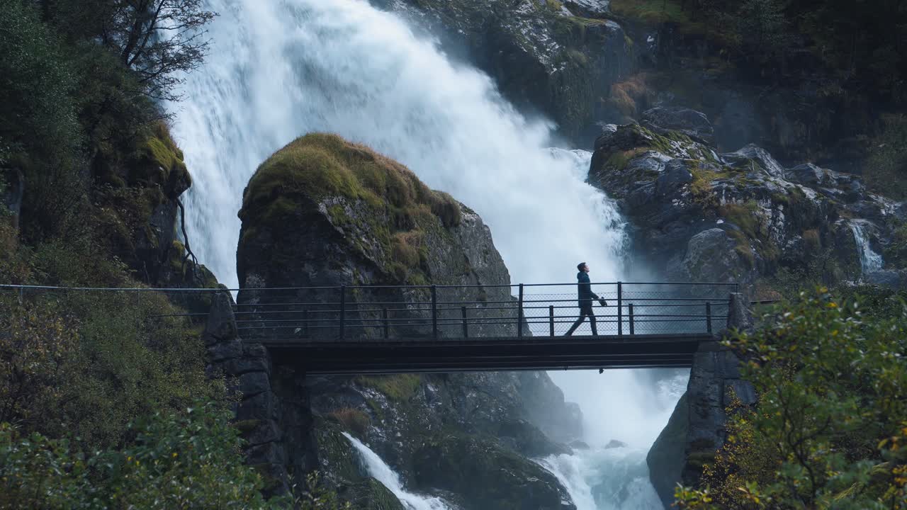 hombre caminando por un pequeño puente con una cascada salvaje en el fondo, noruega briksdalen