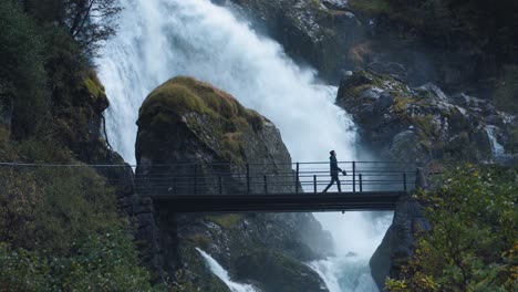 man walking across a small bridge with a wild waterfall on the background, norway briksdalen