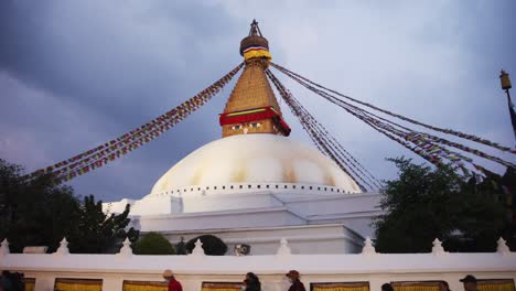 people walk below outside of white and gold temple walls with decorated flags and god symbol on top of round white building