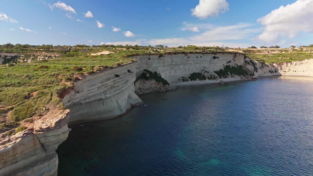 Volo a bassa quota di un drone che sfiora le scogliere calcaree frastagliate di Malta, rivelando le acque cristalline del Mediterraneo e le tradizionali saline costiere scavate nella costa rocciosa