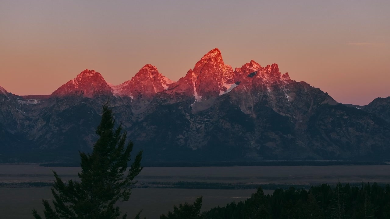 Pink sunrise light glowing on snow covered Grand Teton mountain peaks, aerial telephoto backdrop sunset warmth