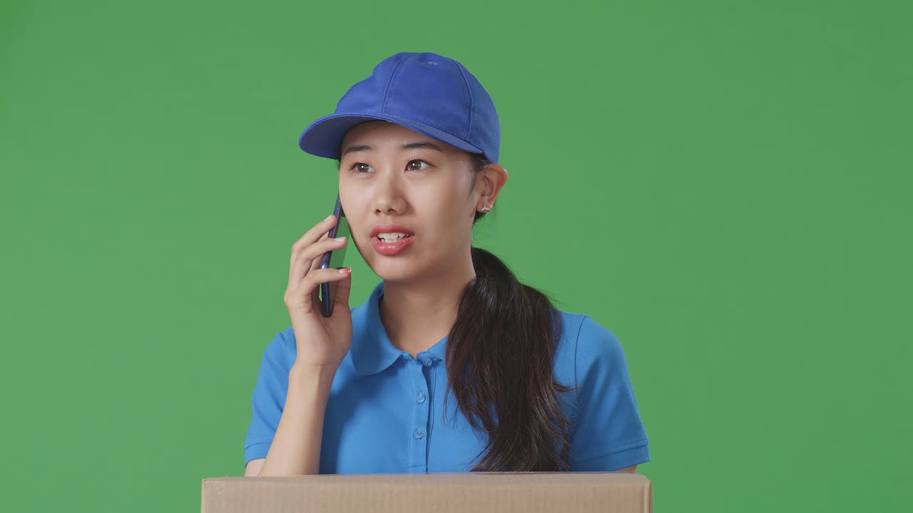 Close Up Of Asian Female Courier In Blue Uniform Talking On Smartphone While Delivering A Carton On Green Screen Background In The Studio