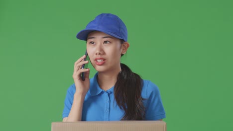 primer plano de una mensajera asiática en uniforme azul hablando en un teléfono inteligente mientras entrega un cartón en el fondo de la pantalla verde en el estudio
