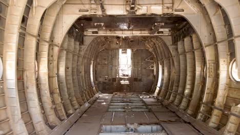 Aerial-zoom-out-drone-shot-of-the-inside-an-old-dismantled-army-plane-monument-placed-on-an-empty-dirt-field,-reviling-a-view-of-trees,-plants-and-a-dirt-trail