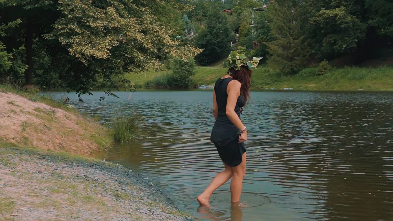 Slim brunette female wearing flower head wreath walking into cold water of lake