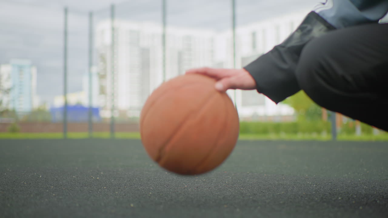 urban outdoor court basketball dribble closeup, crouched player guiding ball with fingertips on textured asphalt, lowangle shot with apartment skyline in background, overcast light, focused training