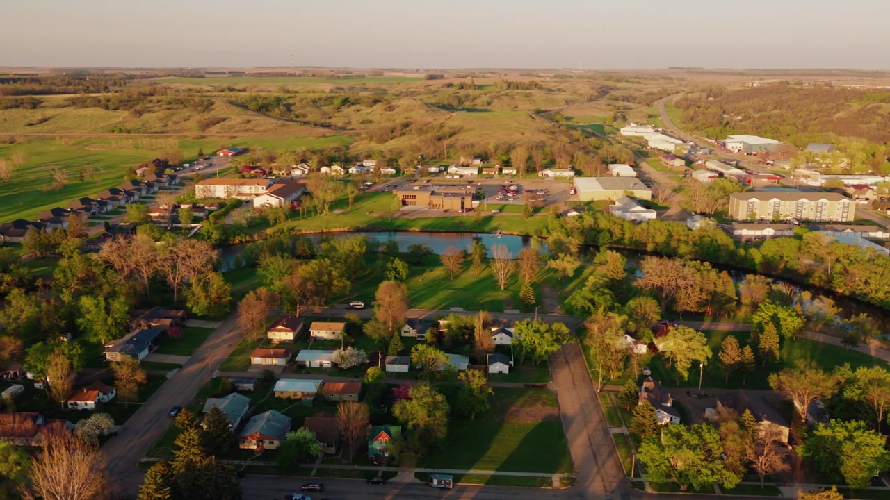 Small town America from above at golden hour: Valley City North Dakota homes, river and prairie hills glowing in warm evening sunlight, aerial drone.