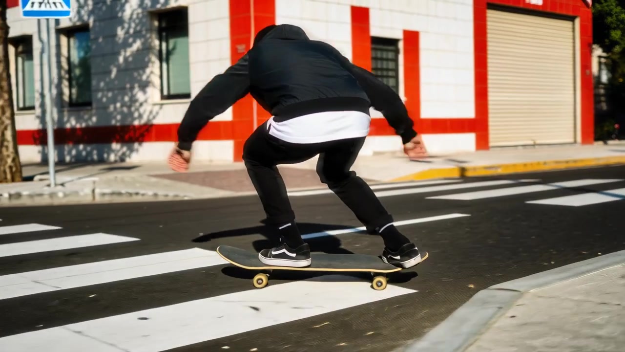Pemain skateboard bertopi kupluk hitam mendorong dengan kaki belakang di atas papan, meluncur melintasi penyeberangan untuk menyeberang jalan.