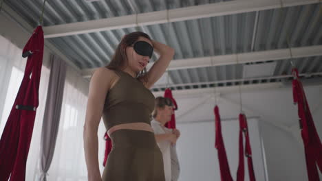 yoga specialists pause momentarily during calming session, one adjusting blindfold while regaining focus in tranquil studio surrounded by suspended red hammocks in peaceful environment