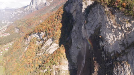 Luftaufnahme-Eines-Wunderschönen-Kleinen-Wasserfalls,-Der-Einen-Regenbogen-Hervorbringt,-Mit-Einem-Bunten-Wald-Im-Herbst-Im-Hintergrund