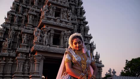 young woman wearing traditional indian clothing and jewelry performing a graceful devotion in front of a temple decorated with statues at sunset