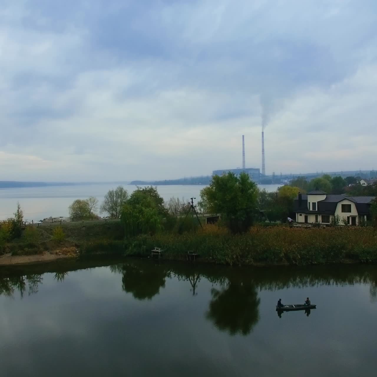Rural area with private houses on the bank of the river. Two people fishing in the boat on the smooth surface of river. Grey cloudy autumn day backdrop