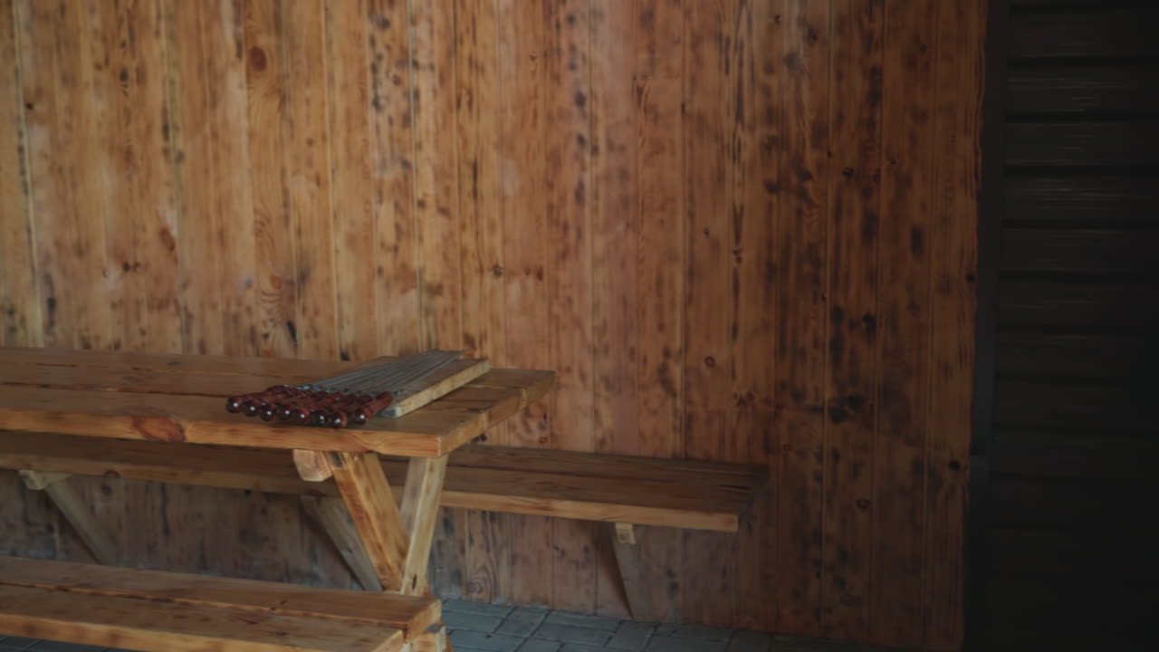Wooden resort seating area features L shaped wooden benches and table with skewers neatly arranged on top, set against textured wood-paneled walls and stone floor