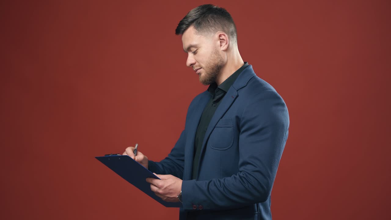 Half-length video of handsome young smiling businessman wearing black shirt and dark blue suit holding the folder taking notes. Dark red background.