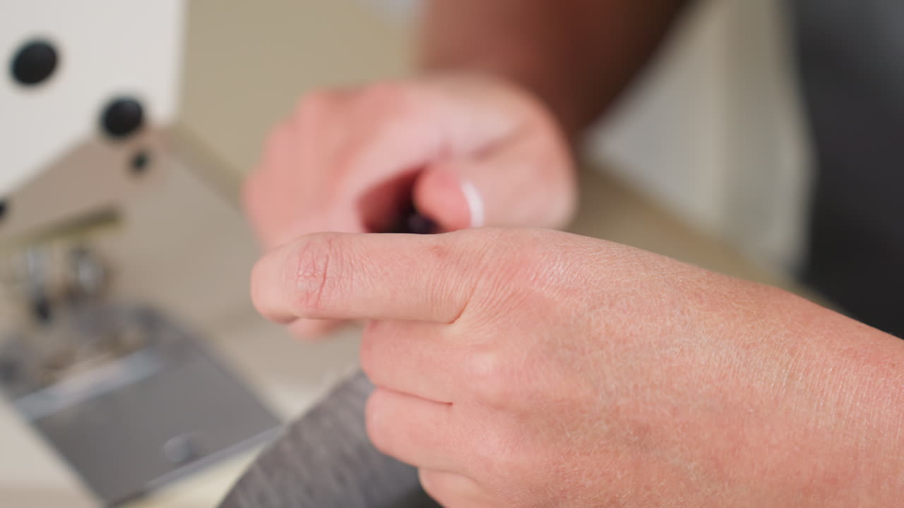 Close up designer hands cutting extra loose threads from fabric edge with small precision scissors focusing on removing imperfections while sewing near machine in brightly lit creative workshop