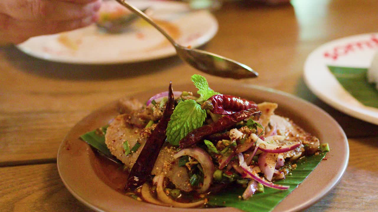 Hand adds garnish to spicy pork salad on banana leaf, natural lighting, close-up perspective