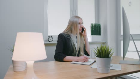 business woman talking on smartphone in office. elegant young blond female in office suit sitting at workplace and negotiating via mobile phone in hand writing down necessary information into notebook