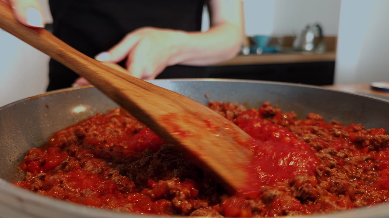 Una persona sala e mescola la salsa bolognese fatta in casa con carne macinata e pomodoro in una padella. Primo piano della preparazione di un classico sugo per la pasta a casa.