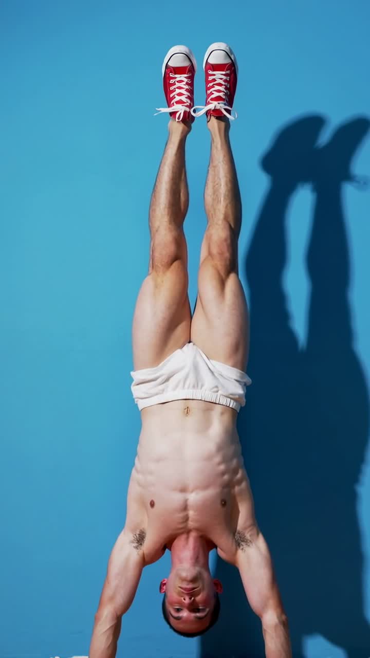 Video verticale: Uomo a torso nudo in equilibrio che esegue una verticale in studio su indicazione per le riprese, scarpe da ginnastica rosse.