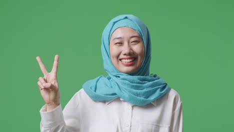 close up of asian muslim woman smiling and showing peace gesture to camera while standing in the green screen background studio