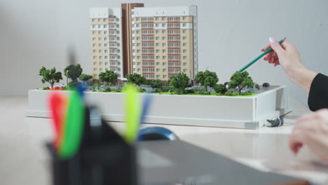 close up of colorful pen holder in foreground with partial view of person pointing pencil at detailed residential building model during architectural explanation