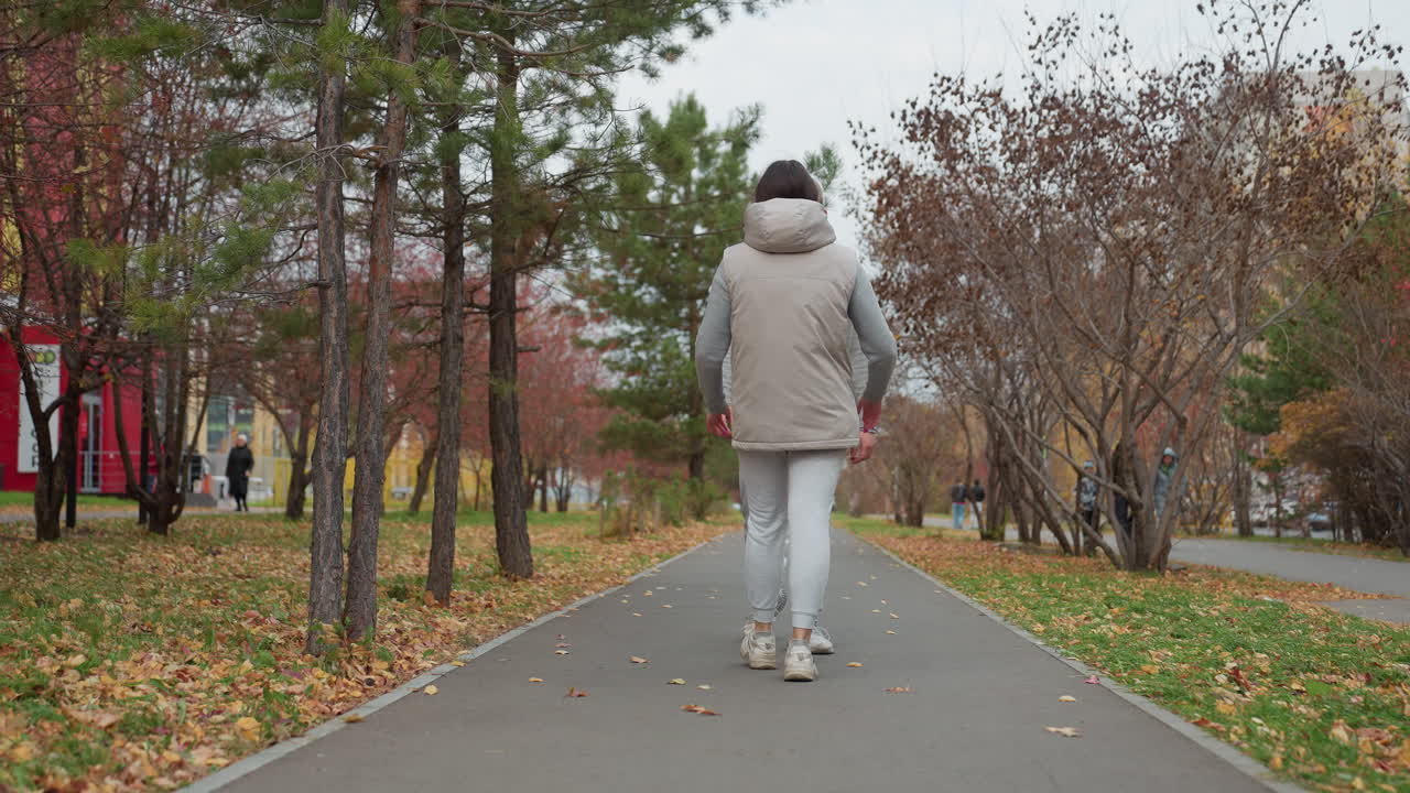 Back view of woman walking along autumn pathway to meet husband as she hugs him tightly under tree-lined avenue with scattered leaves and people strolling in background